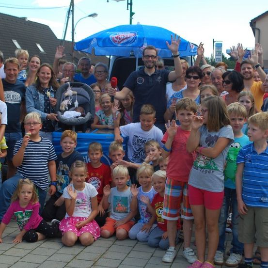Eine Gruppe von Menschen posiert für ein Foto mit einem Mann, der ein Mikrofon hält, und einem Baby in einem Autositz unter einem blauen Regenschirm.