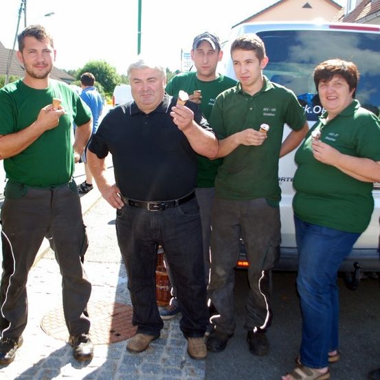 Eine Gruppe von Menschen in grünen Shirts und Hosen steht neben einem weißen Van und hält Eisbecher. Dahinter geht eine Person in einem blauen Shirt. Im Hintergrund befinden sich Häuser und Bäume.