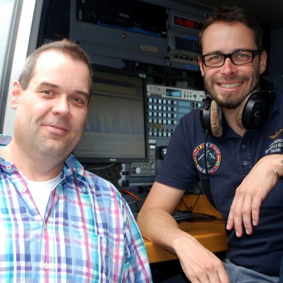 Zwei Männer lächeln und posieren für ein Foto in einem Kontrollraum. Einer trägt eine Brille und Kopfhörer, der andere ein kariertes Hemd. Hinter ihnen sind ein Monitor und verschiedene elektronische Geräte zu sehen.