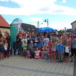 Eine Gruppe von Menschen, einschließlich Erwachsener und Kindern, posiert für ein Foto auf einem Stadtplatz. Ein grüner Banner mit dem Text 'Radio Steiermark' ist sichtbar.