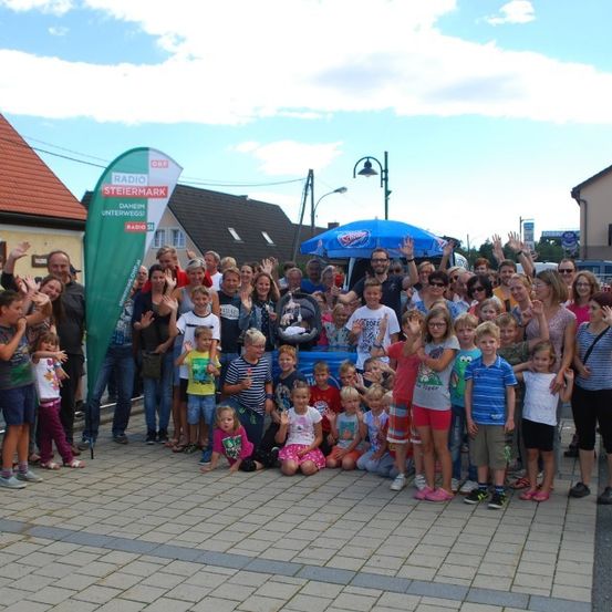 Eine Gruppe von Menschen, einschließlich Erwachsener und Kindern, posiert für ein Foto auf einem Stadtplatz. Ein grüner Banner mit dem Text 'Radio Steiermark' ist sichtbar.