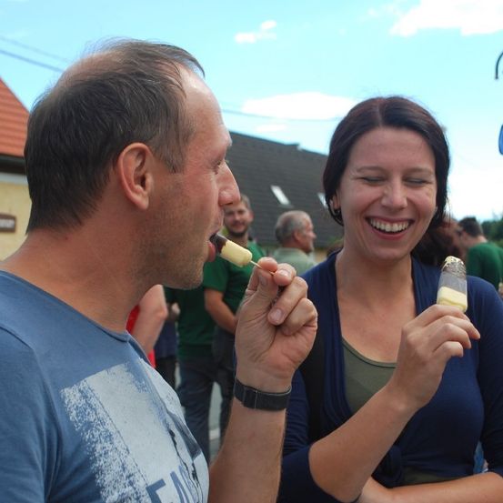 Ein Mann und eine Frau essen Eis am Stiel und lächeln in einer Outdoor-Umgebung, mit Menschen und Gebäuden im Hintergrund.