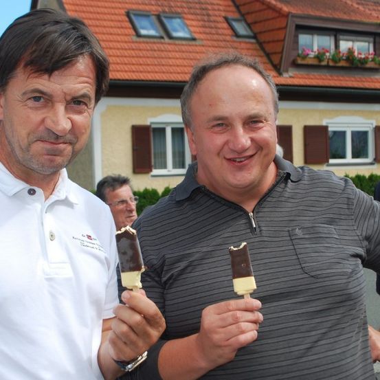 Zwei Männer halten Schokoladen-Eisstangen vor einem Haus mit rotem Dach und Blumen in einer Fensterbank.