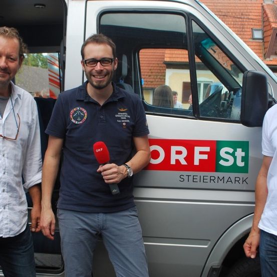 Drei Männer stehen vor einem Van mit ORF Steiermark Aufschrift. Der Mann in der Mitte hält ein Mikrofon.