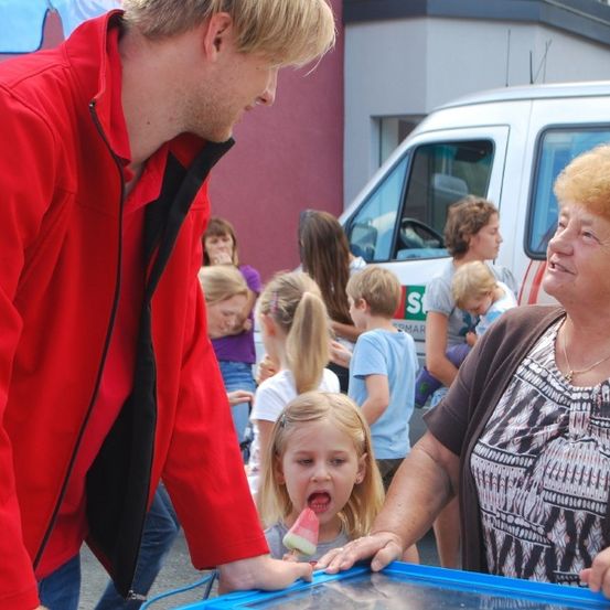 Ein Mann in einem roten Mantel gibt einem kleinen Mädchen ein Eis am Stiel. Eine ältere Frau steht neben ihnen. Andere Kinder und Erwachsene sind im Hintergrund. Ein Van ist in der Nähe geparkt.