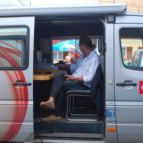 Ein Mann sitzt in einem OFP-Van mit einem Schreibtisch und tippt auf einem Computer. Die Tür des Vans steht offen, und draußen ist ein blauer Regenschirm zu sehen.