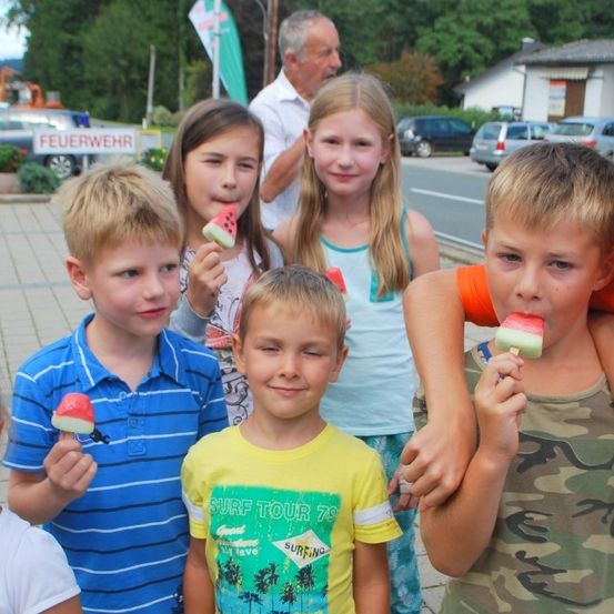 Eine Gruppe von Kindern, die zusammenstehen, jeder einen Wassermelonen-Lollipop hält, einer in einem Tarnhemd, andere in Freizeitkleidung, mit einem Mann im Hintergrund.