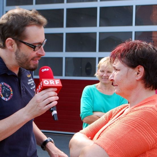 Ein Mann mit einem roten ORF-Mikrofon interviewt eine Frau in Orange, mit zwei anderen Frauen im Hintergrund, eine in Blau und eine in Rosa.