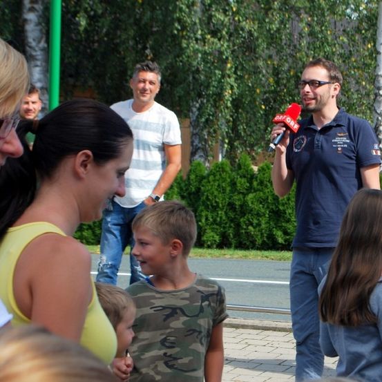 Ein Mann mit Brille spricht in ein Mikrofon, während mehrere Leute zuhören. Es stehen zwei Jungen und zwei Mädchen in der Nähe. Sie sind alle draußen.