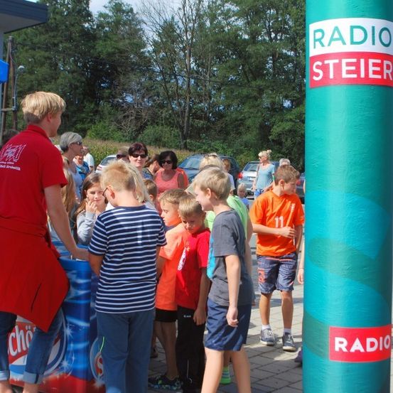 Eine Gruppe von Kindern versammelt sich um einen roten Coca-Cola-Kühler, mit Erwachsenen hinter ihnen, vor einem hohen Radio Steiermark-Banner.