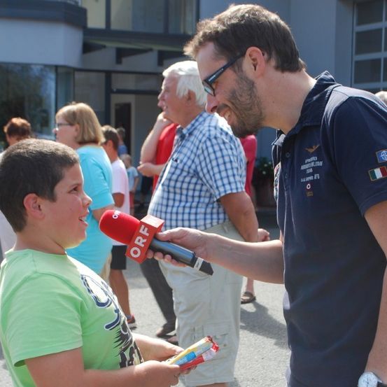 Ein Mann mit Brille interviewt einen Jungen im Freien. Der Junge hält ein rotes Mikrofon mit dem Wort ORF darauf. Hinter ihnen stehen mehrere Menschen und unterhalten sich.