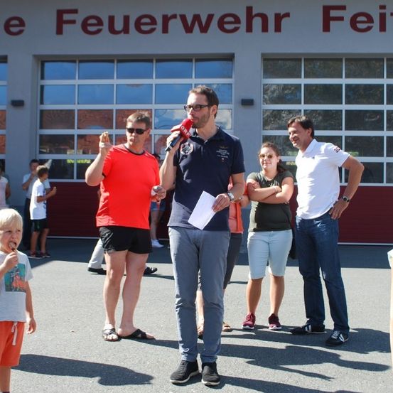 Ein Mann spricht in ein Mikrofon vor einer Menschenmenge an einer Feuerwache. Ein Junge isst ein Eis.