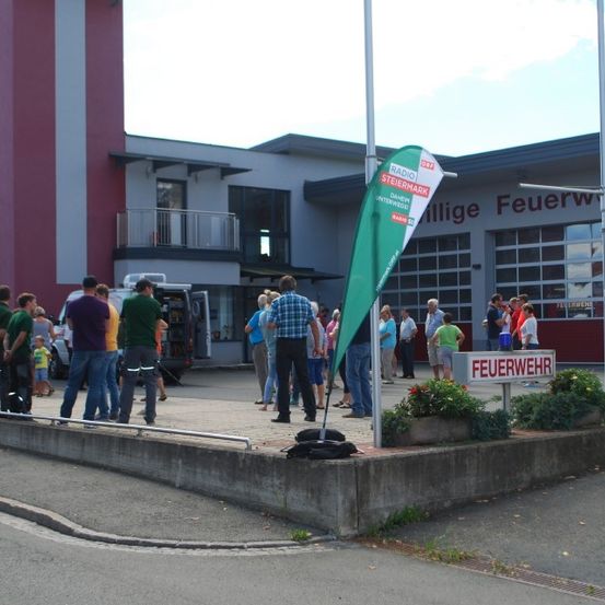 Menschen versammeln sich vor einer Feuerwehrstation mit einem Banner, in der Nähe eines Gebäudes mit Balkon und einem Schild mit der Aufschrift 'Radio Steiermark'.
