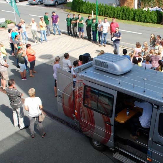 Eine Menschenmenge steht auf einem Bürgersteig und beobachtet einen silbernen Van mit roten Markierungen, einer offenen Tür und einer Person, die mit einem Tisch und einer Flasche davor sitzt.