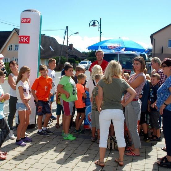 Eine Gruppe von Kindern und Erwachsenen steht auf einem Außenbereich, einige tragen Sandalen und Hausschuhe. Ein Mann hält ein Mikrofon und spricht, wobei sich eine Menschenmenge um ihn herum versammelt. Ein Regenschirm steht in der Nähe, und ein LKW ist am Straßenrand geparkt.