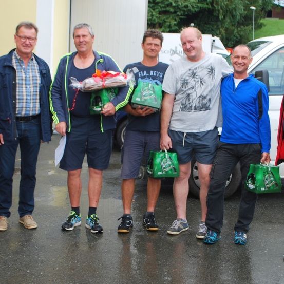 Eine Gruppe von Männern in Freizeitkleidung steht auf einem nassen Gebiet und hält Geschenktüten, mit einem Van und Bäumen im Hintergrund.