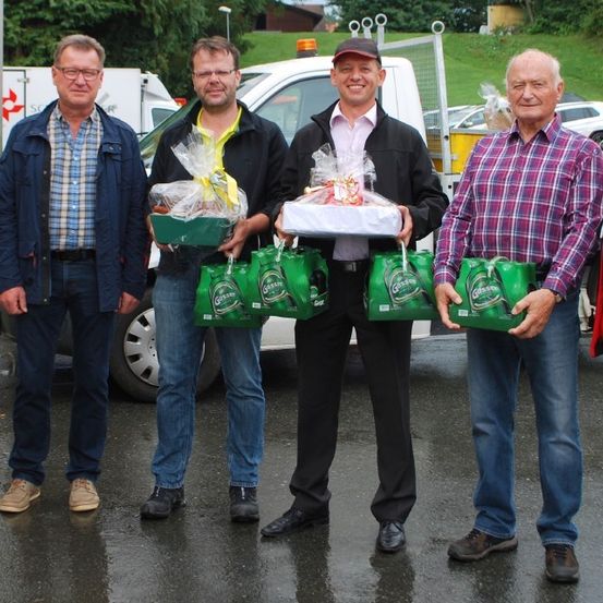 Fünf Männer stehen in einem nassen Bereich und halten Geschenke. Sie tragen Freizeitkleidung. Hinter ihnen stehen Fahrzeuge, einschließlich eines weißen Trucks.