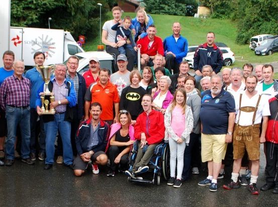 Eine Gruppe von Menschen steht im Freien und lächelt für ein Foto. Sie tragen verschiedene Kleidungsstücke und halten eine Trophäe. Im Hintergrund sind Fahrzeuge geparkt und der Boden ist nass.
