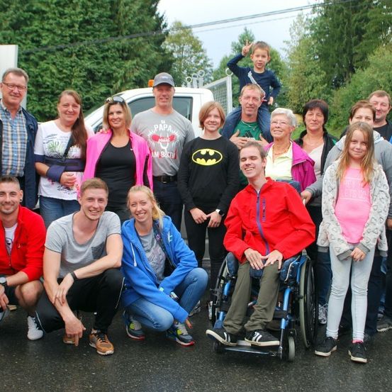 Eine Gruppe von Menschen posiert für ein Foto im Freien. Einige sitzen, andere stehen. Ein Mann im Rollstuhl lächelt. Hinter ihnen steht ein weißer Van.