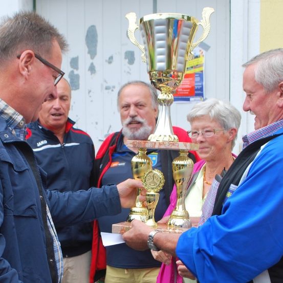 Ein Mann, der einen Pokal hält, lächelt, während ein anderer Mann zusieht. Hinter ihnen lächelt eine Frau mit Brille in die Kamera.