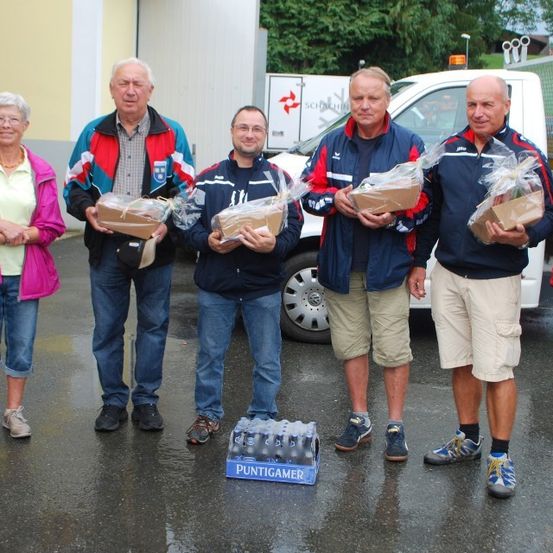 Eine Gruppe von Menschen steht im Regen mit Geschenkboxen. Sie sind lässig gekleidet. Hinter ihnen steht ein weißer LKW mit einem Logo. Ein Karton mit Wasserflaschen liegt auf dem Boden.