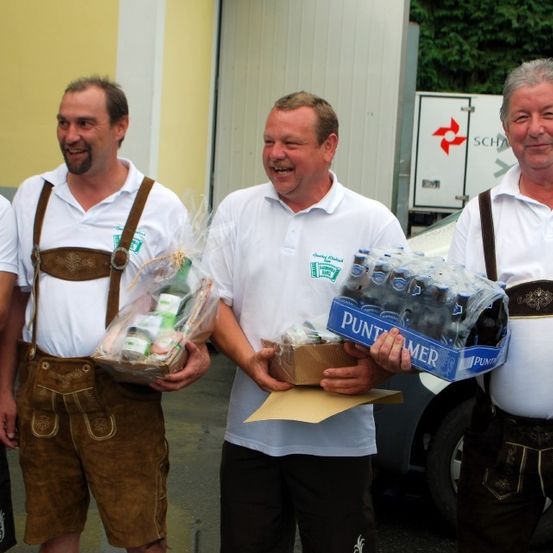 Fünf Männer in traditioneller deutscher Kleidung stehen vor einer Garage. Jeder hält ein Geschenk, einer trägt eine Kiste Bier. Alle tragen weiße Hemden mit grünen Logos und braunen Hosenträgern. Dahinter ist ein weißer Lieferwagen mit einem roten Logo geparkt.