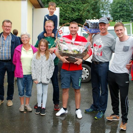 Eine Gruppe von Menschen steht in einem nassen Parkplatz und lächelt. Ein Mann in einem roten Shirt hält einen Korb, während ein anderer in einem grauen Shirt eine Flasche hält. Hinter ihnen ist ein weißer Van geparkt.