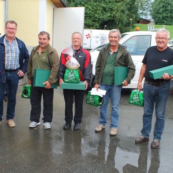 Fünf Männer stehen draußen und halten grüne Geschenkboxen in der Hand, lächeln für ein Foto. Dahinter ist ein Lieferwagen geparkt, und ein Gebäude ist sichtbar.