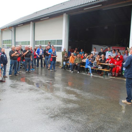 Eine Gruppe von Menschen steht in einer Reihe vor einem Gebäude mit offener Garage. Mehrere Personen sitzen an Tischen mit Flaschen und einem Banner.
