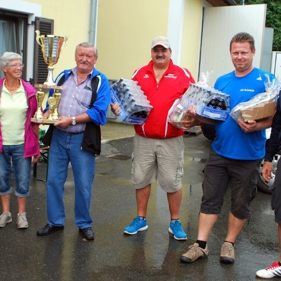 Eine Gruppe von Menschen steht draußen im Regen und hält Trophäen und Pakete. Ein Mann hält eine Trophäe, ein anderer ein Paket, und der Mann in Rot trägt einen Hut. Die Frau trägt eine Brille und eine Halskette. Hinter ihnen steht ein Gebäude mit einem Fenster.