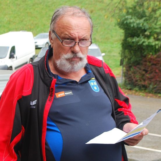 Ein älterer Mann mit Brille, Bart und rot-schwarzer Jacke liest ein Papier auf einem Parkplatz. Hinter ihm sind Fahrzeuge geparkt.