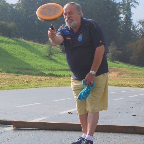 Ein älterer Mann in einem blauen Poloshirt und gelben Shorts steht auf einer nassen Straße und hält eine Frisbee. Die Umgebung umfasst einen Grashügel und Bäume.