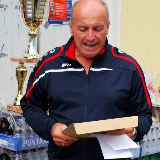 Ein älterer Mann in einem dunkelblauen Jacke mit roten und weißen Streifen liest ein Papier und lächelt, mit einem Pokal und Flaschengetränken im Hintergrund.