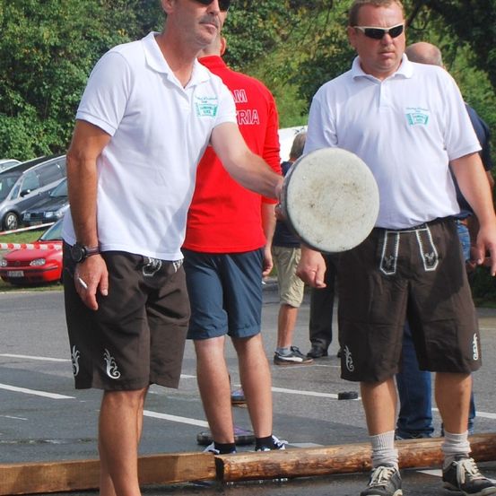 Ein Mann hält eine Frisbee, während ein anderer Mann neben ihm steht. Sie tragen beide weiße Poloshirts und braune Shorts. Hinter ihnen stehen Leute, und im Hintergrund sind Autos geparkt.