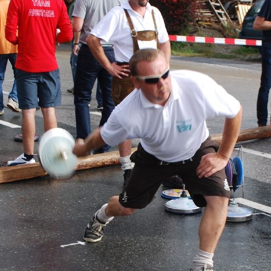 Ein Mann mit Sonnenbrille und weißem Shirt wirft einen Frisbee auf einer Straße, hinter ihm stehen mehrere Menschen in Freizeitkleidung und Turnschuhen.