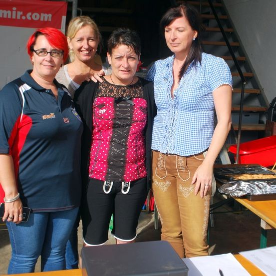 Vier Frauen stehen zusammen, zwei mit Brille, eine mit roten Haaren. Sie sind in der Nähe eines Tisches mit Essen, Treppen und einem Banner im Hintergrund.