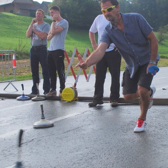 Vier Männer sind draußen auf einer nassen Straße. Ein Mann wirft einen Ball, während drei Männer zuschauen. Ein gelbes Objekt steht auf der nassen Straße.