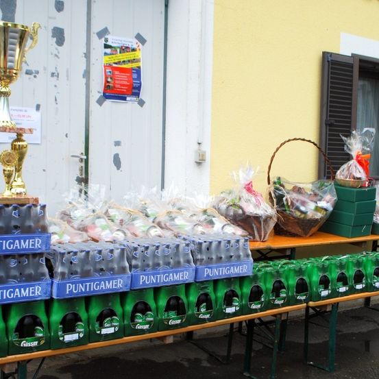 Ein Tisch mit verschiedenen Artikeln zum Verkauf, einschließlich eines Pokals, Bierflaschen und verpackten Geschenken. Der Tisch steht neben einer gelben Wand mit einer geschlossenen Tür und einem Plakat.