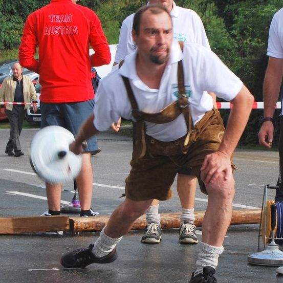 Ein Mann in traditioneller deutscher Kleidung wirft einen Frisbee bei einem Wettkampf im Freien. Dahinter steht ein Mann in einem roten Pullover mit dem Wort Österreich. In der Nähe beobachten andere Leute das Ereignis.