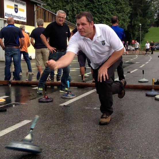 Ein Mann in einem weißen Shirt wirft einen Hammer im Freien, während andere zusehen, mit mehreren Hämmern und einem Holzstamm auf dem Boden.
