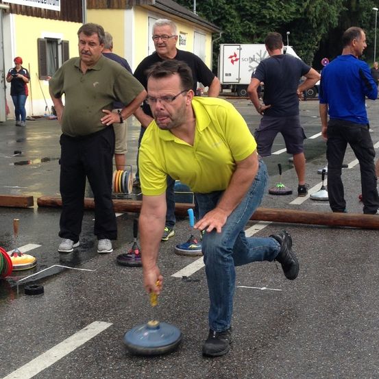 Eine Gruppe von Männern spielt ein Spiel auf einer nassen Straße. Ein Mann in einem gelben Shirt beugt sich nach vorne und hält ein blaues Objekt. Andere Männer stehen hinter ihm und schauen zu.