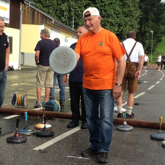 Ein älterer Mann hält eine Scheibe in einem Park. Mehrere Leute stehen hinter ihm und schauen zu. Bunte Objekte und eine große Rohr liegen auf dem Boden. Bäume und ein Gebäude sind im Hintergrund.