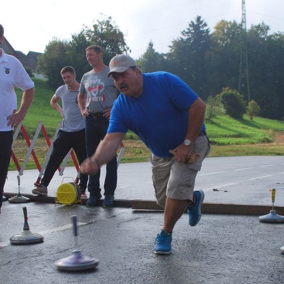 Mehrere Männer spielen ein Spiel auf einer nassen Oberfläche. Ein Mann in Blau läuft, ein Mann in Grau steht und ein Mann in Weiß steht hinter ihnen.