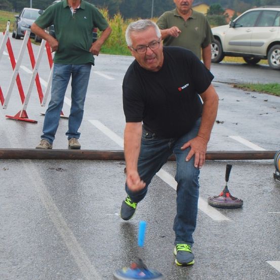 Ein älterer Mann wirft einen Dart auf ein Ziel, während zwei andere zuschauen. Sie befinden sich auf einer nassen Straße mit Verkehrskegeln und geparkten Autos.