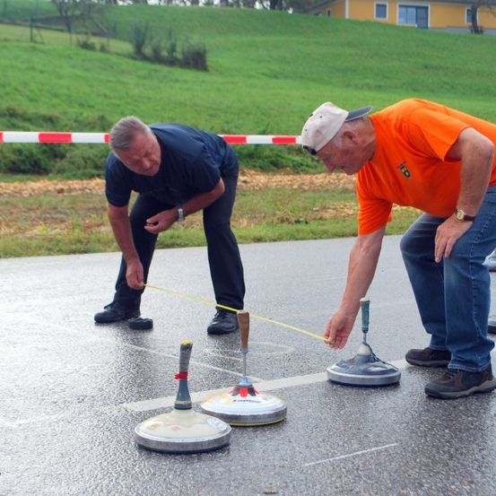 Zwei Männer sind in einem Outdoor-Spiel engagiert und benutzen ein gelbes Maßband, um Entfernungen auf einem nassen, markierten Boden zu messen. Sie bücken sich beide, einer schaut auf das Maßband und der andere lächelt.