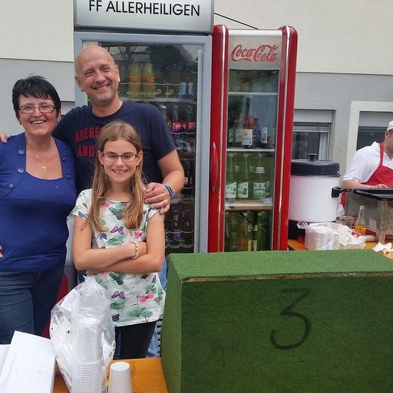 Drei Personen stehen vor einem Coca-Cola-Kühler, mit einer grünen Box mit der Aufschrift '3' im Vordergrund. Ein Mann in einer weißen Schürze steht hinter dem Kühler.