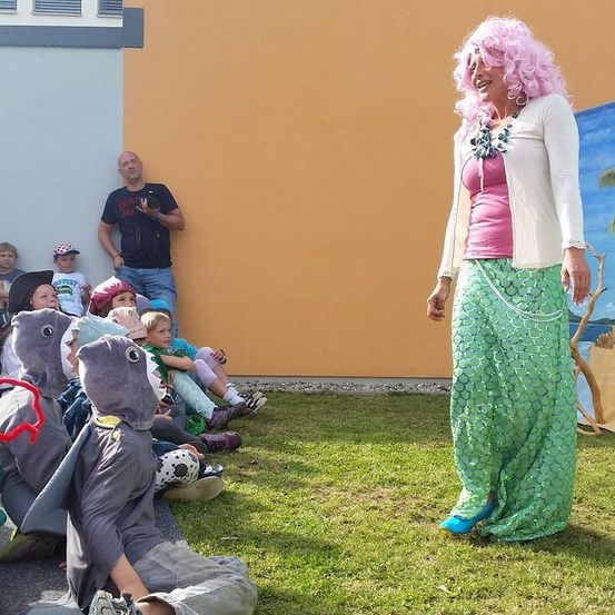 Eine Frau, die als Meerjungfrau verkleidet ist, unterhält eine Gruppe von Kindern in Haikostümen. Ein Mann mit einer Kamera steht hinter ihnen und sie befinden sich in der Nähe einer bemalten Wand.