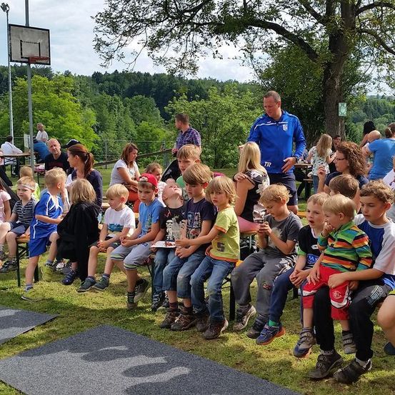 Eine Gruppe von Kindern sitzt auf einer Bank in einem Park, mit Erwachsenen in der Nähe. Ein Mann in einem blauen Jackett spricht die Kinder an. Im Hintergrund befindet sich ein Basketballkorb und ein Gebäude.