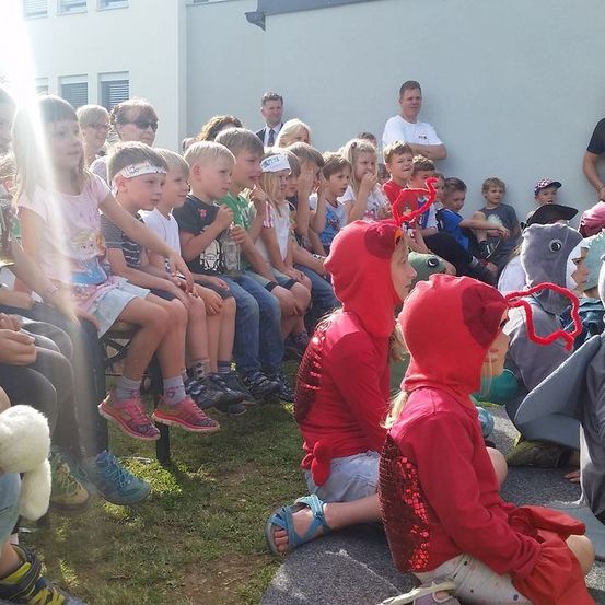 Eine Gruppe von Kindern sitzt auf dem Gras draußen und trägt rote Kostüme. Einige halten Flaschen. Dahinter sitzen und stehen einige Erwachsene, und ein Mann hält eine Kamera. Die Kinder schauen sich wahrscheinlich eine Aufführung an.