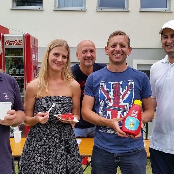 Eine Gruppe von Menschen posiert für ein Foto im Freien. Einer hält ein Getränk und einen Teller mit Essen, ein anderer hält eine Flasche Ketchup, während andere Tassen halten. Im Hintergrund befindet sich ein Coca-Cola-Automat und ein Gebäude.